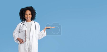 Photo for Happy young african american curly woman doctor therapist in white coat hold free space in hand and tablet isolated on blue background, studio, panorama. Ad, offer, medicine service, health care - Royalty Free Image