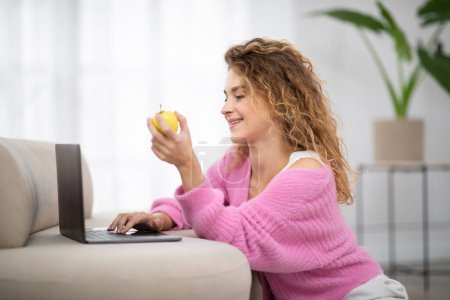 Foto de Mujer milenaria sonriente relajarse con el ordenador portátil en casa y comer manzana, Feliz hermosa hembra navegar por Internet en el ordenador mientras descansa en el interior de la sala de estar, Disfrutando del ocio doméstico - Imagen libre de derechos