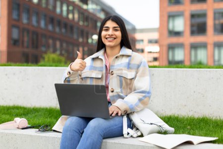 Photo for Happy hispanic student lady sitting with laptop outdoors, gesturing thumbs up and smiling, approving distance online education, having remote lesson in campus park. I like e-learning - Royalty Free Image