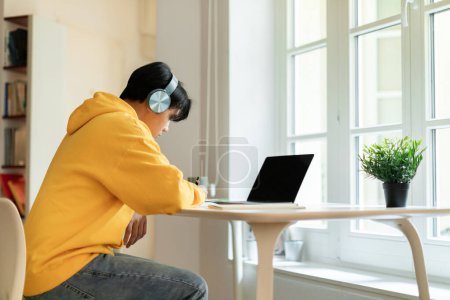 Photo for Online Education Concept. Profile view of asian teen guy taking notes during webinar on laptop, studying online at home. Boy writing in his copybook doing homework. Education and remote classes - Royalty Free Image
