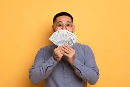 Photo for Big Profit. Excited Young Asian Guy Holding Dollar Cash Fan Near Face, Happy Millennial Man With Money In Hands Standing Over Yellow Studio Background, Celebrating Success, Copy Space - Royalty Free Image