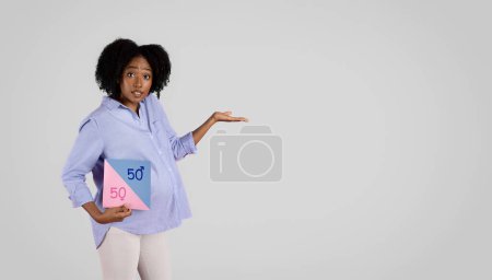 Téléchargez les photos : Confus millénariste afro-américaine frisée femme enceinte avec grand ventre tenir bannière avec 50 pour cent, pointez la main sur l'espace vide, isolé sur fond de studio gris. Sexe fête, garçon ou fille attendre - en image libre de droit