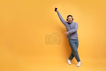 Téléchargez les photos : Amusant fou. gai asiatique homme dans sans fil casque dansant sur jaune fond, heureux jeune homme tenant Smartphone moderne dans la main, tromper dans studio, pleine longueur prise de vue, Copier l'espace - en image libre de droit