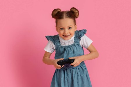 Foto de Emocionada niña jugando videojuegos con Joystick mientras está de pie sobre el fondo del estudio rosa, Feliz niña preadolescente divirtiéndose, Disfrutando de los aparatos modernos para el ocio, Copiar el espacio - Imagen libre de derechos