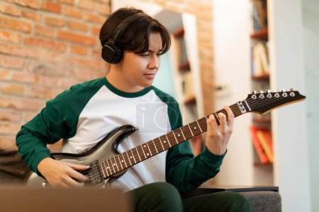 Foto de Joven músico. Korean Teen Boy Disfrutando del ritmo mientras toca la guitarra eléctrica, usando auriculares sentados cerca de la computadora portátil en interiores. Joven estudiante chico aprender a tocar instrumento musical en casa - Imagen libre de derechos