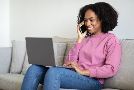 Photo for Cheerful millennial black woman typing on computer, reads message, calls by phone, sits on sofa in living room interior. Video call, communication remotely, meeting, work and study at home - Royalty Free Image