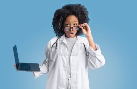Photo for Shocked young african american curly lady doctor therapist in white coat with laptop takes off glasses, isolated on blue background, studio. Surprise, good news, medicine service, health care - Royalty Free Image