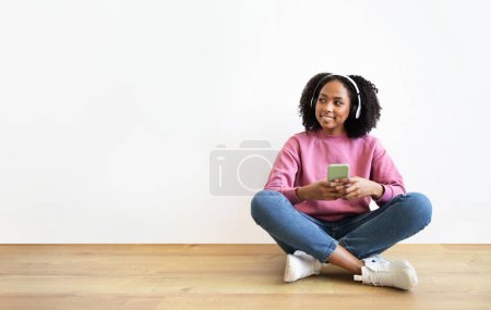 Téléchargez les photos : Heureuse femme noire millénaire dans les écouteurs sans fil assis sur le sol, bavarder sur smartphone, écouter de la musique, regarder l'espace vide à l'intérieur de la chambre sur fond de mur blanc. Réseaux sociaux, application audio - en image libre de droit