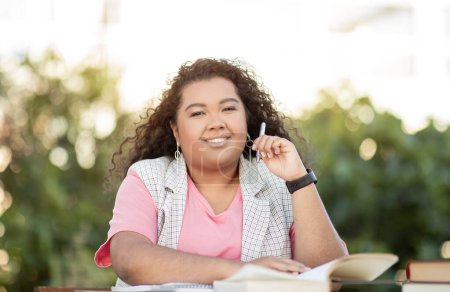 Téléchargez les photos : Portrait de heureuse étudiante hispanique apprenant assis à l'extérieur. Apprenante dame lit des livres et prend des notes posant avec stylo et manuels, faire des devoirs de collège dans le parc à l'extérieur - en image libre de droit