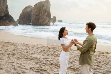 Photo for Loving couple holding hands and looking at each other, having romantic date on the beach, standing near ocean shore, free space. A couples romantic moment on the coast - Royalty Free Image