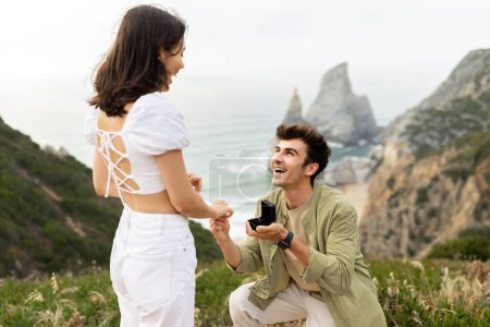 Foto de Serenata oceánica. Joven hombre feliz haciendo propuesta a su novia en la hermosa vista de la costa. Fecha junto al mar y romance especial matrimonio al aire libre - Imagen libre de derechos
