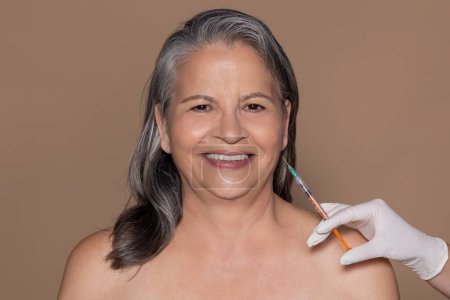 Foto de Sonriente mujer europea de pelo gris senior disfrutar de procedimiento de belleza, cosmetólogo hace inyección de arrugas, aislado en fondo de estudio beige. Cuidado de la piel, botox, cosmetología - Imagen libre de derechos