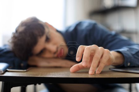 Téléchargez les photos : Burnout professionnel et dépression. Arabe jeune homme épuisé de surmenage, assis triste tête reposante sur la table dans le bureau moderne, foyer sélectif. Homme déprimé souffrant d'apathie Problème - en image libre de droit