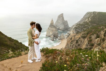 Photo for Loving young couple embracing, having romantic date and enjoying breathtaking view at coastline, standing at the rocks near ocean shore, full length, free space - Royalty Free Image