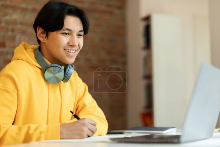 Téléchargez les photos : Gai chinois adolescent guy écriture dans notebook pendant webinaire ou leçon sur ordinateur portable, assis au bureau à la maison. Asiatique adolescent ayant conférence en ligne, prendre des notes souriant à l'ordinateur. Vue latérale - en image libre de droit