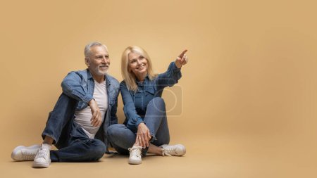 Téléchargez les photos : Heureux couple de personnes âgées positif homme aux cheveux gris et femme blonde portant tenue décontractée assis ensemble sur le sol sur fond beige, pointant vers l'espace de copie, montrant belle affaire, panorama - en image libre de droit