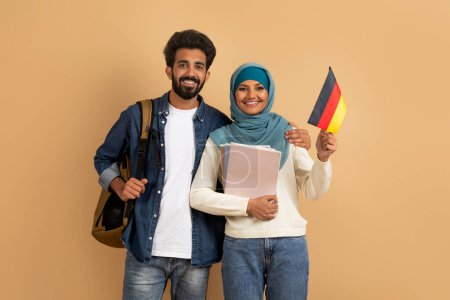 Téléchargez les photos : Éducation internationale. Couple étudiant arabe avec sacs à dos tenant le drapeau allemand, portrait d'un homme et d'une femme musulmans souriants dans le hijab avec des classeurs posant sur fond de studio beige, espace de copie - en image libre de droit