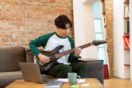 Photo for Japanese Teenager Boy Mastering Electric Guitar Chords, Using Computer And Internet Technology for Music Education, Having Online Lesson At Home. Boy Learning To Play Favorite Songs Near Laptop - Royalty Free Image