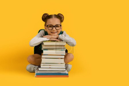 Photo for Back To School Concept. Cute Little Schoolgirl Leaning At Book Stack And Smiling To Camera, Happy Preteen Female Child In Eyeglasses Posing Isolated Over Yellow Studio Background, Copy Space - Royalty Free Image