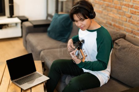 Téléchargez les photos : Éducation musicale numérique. Japanese Teen Boy utilise un ordinateur portable pour des leçons de guitare en ligne, immergé dans la musique avec écouteurs, accords de jeu sur instrument électrique, assis à la maison. Loisir créatif et talent - en image libre de droit