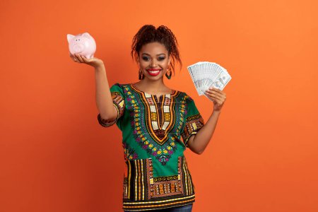 Foto de Ahorros, depósitos bancarios, inversiones. Mujer negra bonita y feliz vistiendo ropa africana sosteniendo alcancía rosa y un montón de billetes de dinero en efectivo, fondo naranja - Imagen libre de derechos