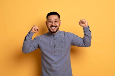 Photo for Big Luck. Overjoyed Young Asian Man Celebrating Success With Raised Fists, Euphoric Happy Millennial Guy In Eyeglasses Cheering Win, Standing Over Yellow Studio Background, Copy Space - Royalty Free Image