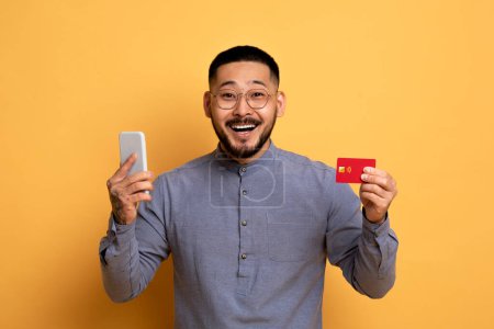 Photo for E-Commerce Concept. Excited Young Asian Man Holding Smartphone And Credit Card, Happy Millennial Male Making Internet Shopping Or Online Payments While Standing Over Yellow Background, Copy Space - Royalty Free Image