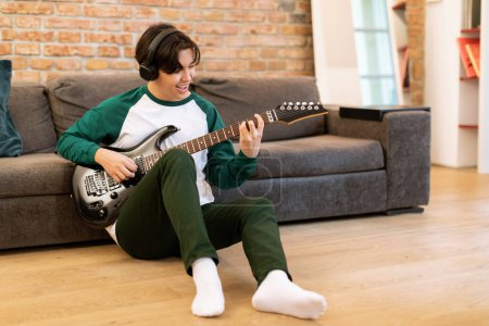 Foto de Educación musical y ocio. Joven músico japonés aprendiendo a tocar, practicando guitarra eléctrica sentada en el piso en el interior de la sala de estar moderna, usando auriculares inalámbricos - Imagen libre de derechos