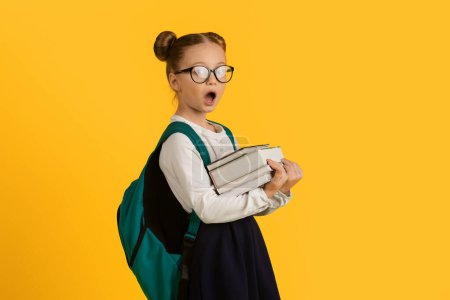 Photo for Shocked Cute Little Schoolgirl Wearing Eyeglasses Holding Stack Of Books And Looking At Camera, Cute Female Pupil Standing With Open Mouth, Having Too Much Homework, Yellow Background, Copy Space - Royalty Free Image