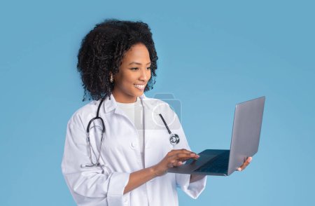 Photo for Happy young african american woman doctor therapist in white coat chatting on laptop, isolated on blue background, studio. Video call with patient, medicine service, health care, treatment remotely - Royalty Free Image