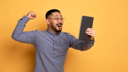 Téléchargez les photos : Heureux jeune homme asiatique avec tablette numérique célébrant le succès, regardant l'écran du pad et secouer le poing, chanceux millénariste gars jeu en ligne, en utilisant la plus récente application mobile sur fond jaune, espace de copie - en image libre de droit