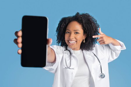 Photo for Glad young african american curly woman doctor in white coat point finger at smartphone with empty screen isolated on blue background, studio. App, blog, website, medicine service, health care - Royalty Free Image