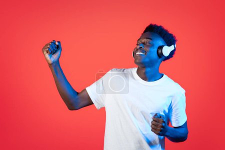 Foto de Feliz joven afroamericano guapo alegre con camiseta blanca usando auriculares estéreo inalámbricos modernos, escuchando música con los ojos cerrados y bailando sobre fondo rojo en luz de neón - Imagen libre de derechos