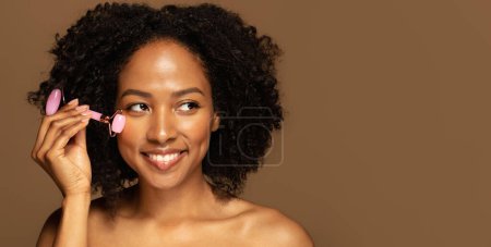 Téléchargez les photos : Belle jeune femme afro-américaine à moitié nue faisant un massage du visage avec un rouleau de jade dans la routine de soins du visage du matin, regardant l'espace de copie et souriant, gros plan, fond de studio, bannière - en image libre de droit