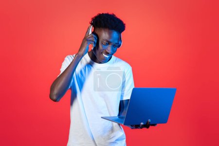 Téléchargez les photos : Joyeux jeune homme noir portant un t-shirt blanc utilisant des écouteurs sans fil modernes et un ordinateur portable PC sur fond rouge dans la lumière au néon, DJ travaillant sur ordinateur, mixant des pistes de musique, espace de copie - en image libre de droit