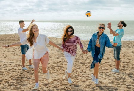 Téléchargez les photos : Heureuses femmes millénaires dansent comme l'équipe de pom-pom girls, les hommes européens et arabes jouent au match de balle, volley-ball, profiter week-end sur la plage, en plein air. Vacances en été, le sport et le plaisir à temps libre - en image libre de droit