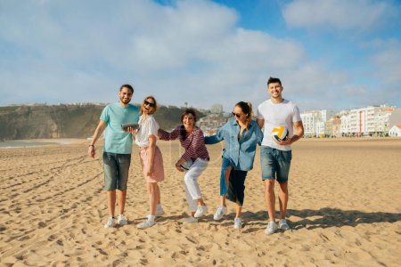 Foto de Sonriendo milenario árabe y europeo personas en casual con pelota se divierten en la playa en fin de semana, al aire libre. Picnic con amigos, vacaciones y caminar juntos, anuncio y oferta, vacaciones - Imagen libre de derechos