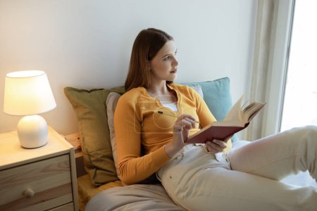 Téléchargez les photos : Loisirs pour ados. Jeune femme blonde lisant et apprenant avec le livre, prenant des notes allongées dans la chambre à coucher à l'intérieur confortable de la maison. Étudiant lit livre de poche sur le week-end, Poser regardant à l'extérieur - en image libre de droit