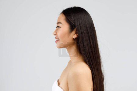 Téléchargez les photos : Portrait de profil de heureuse belle femme asiatique à la peau parfaite et aux cheveux épais, regardant de côté et souriant, posant sur fond gris, espace de copie - en image libre de droit