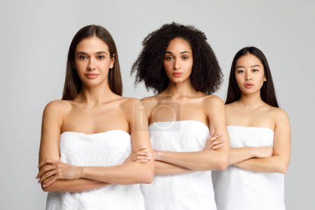 Téléchargez les photos : Belles femmes noires, caucasiennes et asiatiques posant avec les bras croisés en serviettes de bain blanches, debout en ligne sur fond gris, plan studio, bannière - en image libre de droit