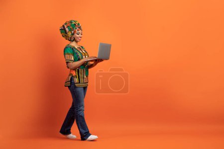 Foto de Trabajo remoto. Sonriendo positiva bastante millennial africana americana mujer vistiendo traje nacional freelancer digital nómada caminar con el ordenador portátil sobre fondo naranja, longitud completa, espacio de copia - Imagen libre de derechos