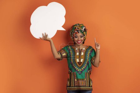 Téléchargez les photos : Inspiré jeune femme noire joyeuse en costume africain avec bulle de conversation vierge sur sa tête sur fond de studio orange, pointant du doigt, eureka, espace pour le design, dame ont de belles idées - en image libre de droit