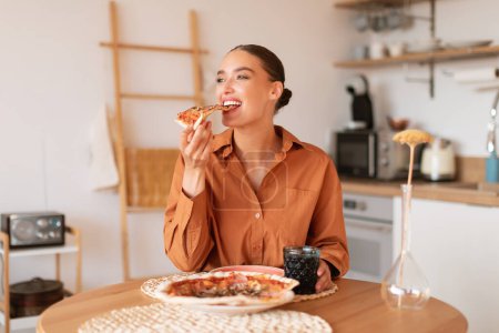 Téléchargez les photos : Une part de bonheur. Heureuse jeune femme caucasienne affamée assise à table dans la cuisine et mangeant de savoureuses pizzas maison, appréciant et souriant, espace libre - en image libre de droit