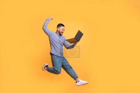 Téléchargez les photos : Émotionnel jeune homme asiatique sautant avec ordinateur portable à la main, joyeux homme millénaire regardant l'écran d'ordinateur et secouant le poing, célébrant la victoire en ligne, s'amusant sur fond de studio jaune, espace de copie - en image libre de droit