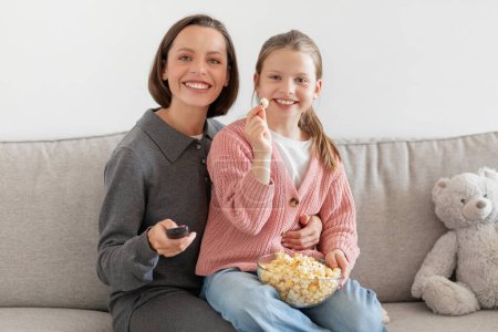 Photo for Positive millennial caucasian woman and teenager daughter eating popcorn, watching tv on sofa in living room interior. Entertainment, fun and movie evening with snacks, rest at home - Royalty Free Image