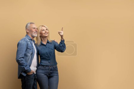 Photo for Beautiful cheerful blonde senior woman showing her excited handsome grey-haired bearded husband nice offer, pointing at copy space, old couple isolated on beige studio background - Royalty Free Image