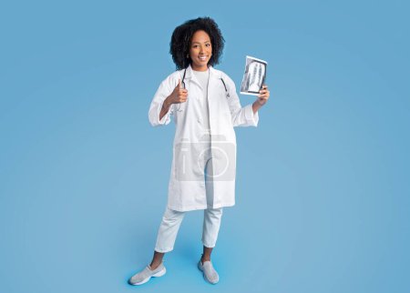 Photo for Happy young african american curly woman doctor therapist in white coat make thumb up sign, show x-ray picture, isolated on blue background, studio. Good result of treatment, medicine, health care - Royalty Free Image