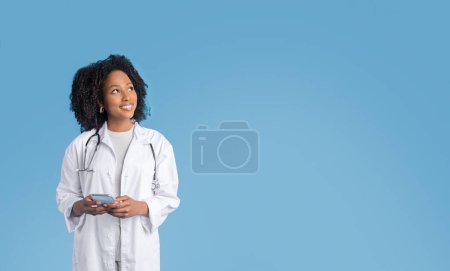 Téléchargez les photos : Joyeux jeune afro-américaine bouclée dame médecin thérapeute en manteau blanc utiliser téléphone, regarder l'espace libre, isolé sur fond bleu, studio. Annonce et offre, service de médecine, soins de santé à distance - en image libre de droit