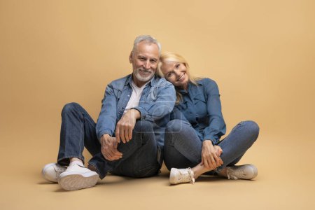 Téléchargez les photos : Aimant beau couple heureux et joyeux homme aux cheveux gris et femme blonde assis sur le sol, fond de studio beige, câlins, souriant à la caméra, portant une tenue décontractée en denim. Portrait de famille - en image libre de droit