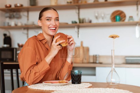 Foto de Saborear lo simple. Mujer feliz comiendo hamburguesa comida rápida de entrega, sentado en la mesa en la cocina y sonriendo a la cámara, disfrutando de la comida rápida, espacio libre. Hambre de carbohidratos - Imagen libre de derechos